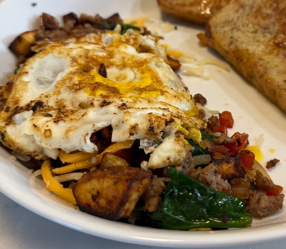 A white bowl of sweet potato and sausage hash with red pepper and wilted spinach under a crisp-edged fried egg and shredded cheese, the corner of a slice of toast behind it.