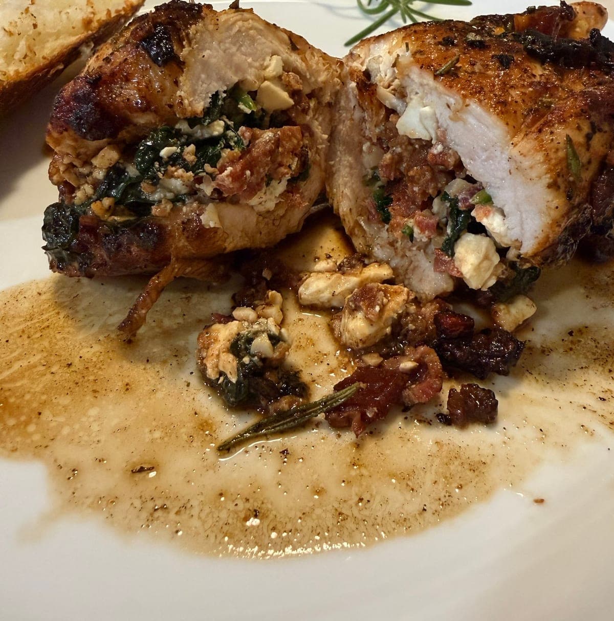 Two halves of a bacon and feta stuffed chicken breast on a white plate, cut open to show the spinach, crumbled feta and chopped bacon packed inside a dark, well-seasoned crust, sitting in a pool of the rosemary butter pan sauce with a rosemary sprig and the edge of a baked potato alongside.