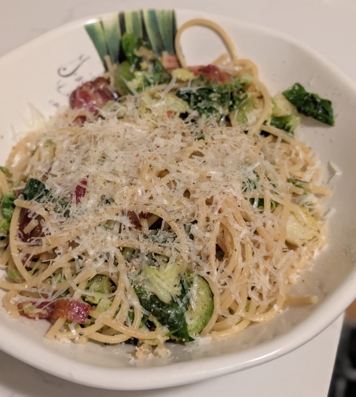A white bowl with a painted green leaf pattern holding spaghetti tossed with halved brussels sprouts, wilted leaves and pieces of bacon, under a heavy shower of grated parmesan flecked with black pepper and red pepper flakes.