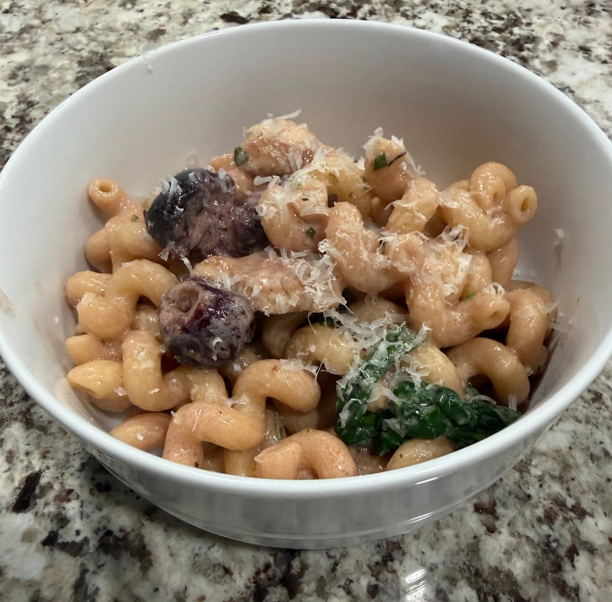 A white bowl of corkscrew pasta in a pink mascarpone sauce with pieces of chicken, two dark cherries, wilted spinach, flecks of herbs and grated parmesan on top.