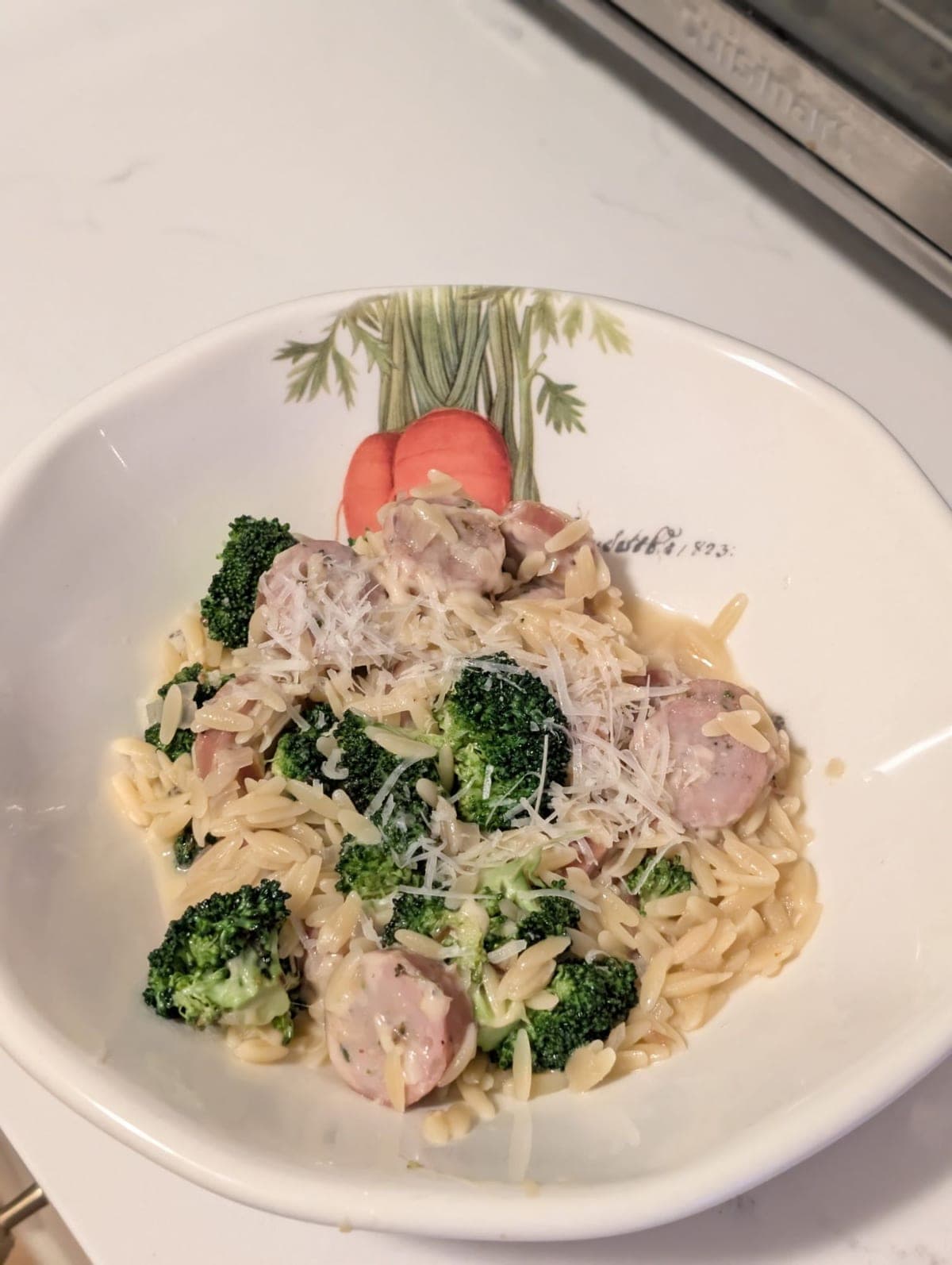 Chicken sausage, broccoli and orzo in a shallow white bowl painted with a carrot — browned sausage coins and broccoli florets folded through creamy orzo, under a heap of coarsely shredded parmesan.