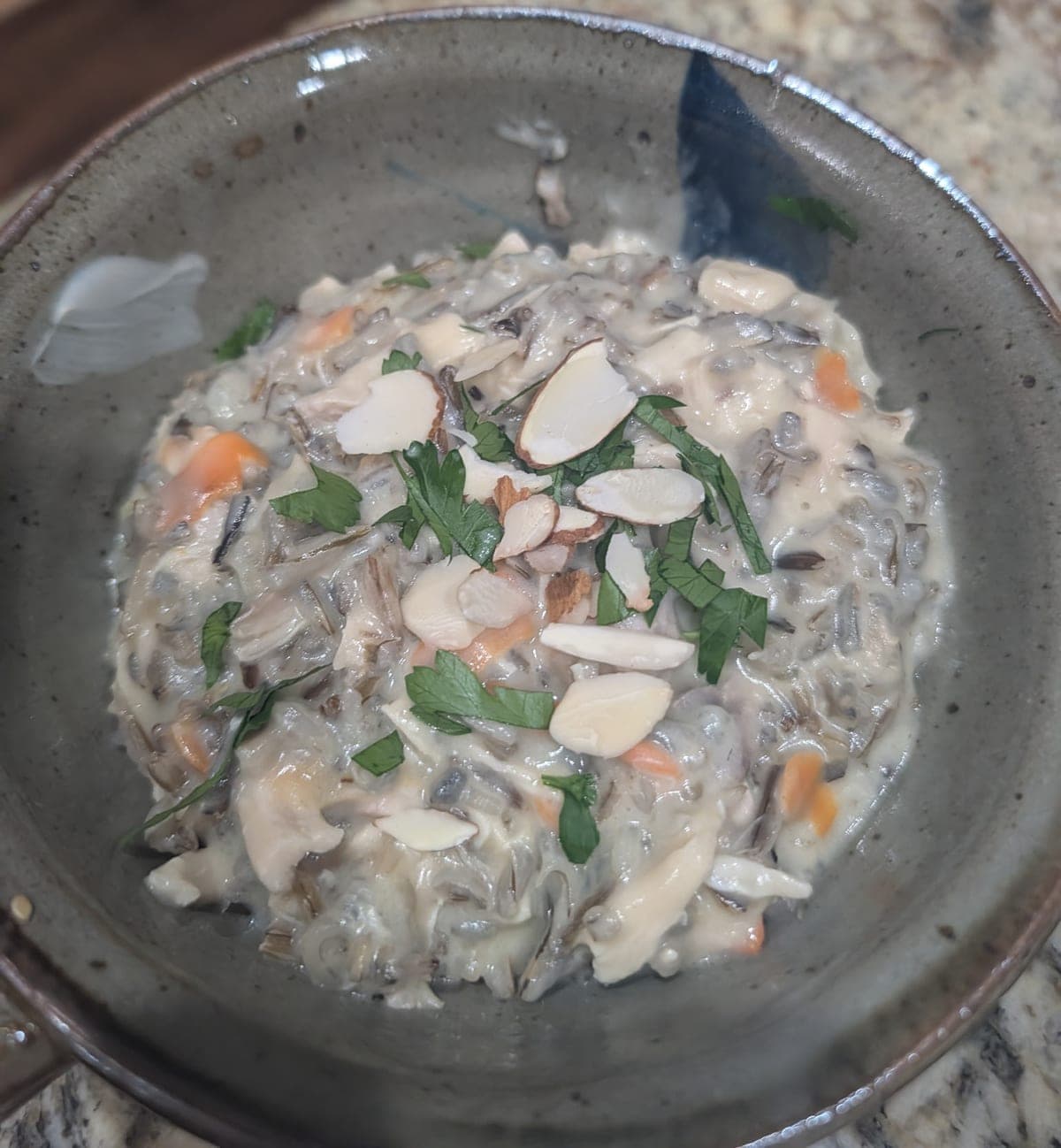 A speckled stoneware bowl of thick, creamy wild rice soup with shredded chicken and bits of carrot, topped with sliced almonds and chopped parsley.