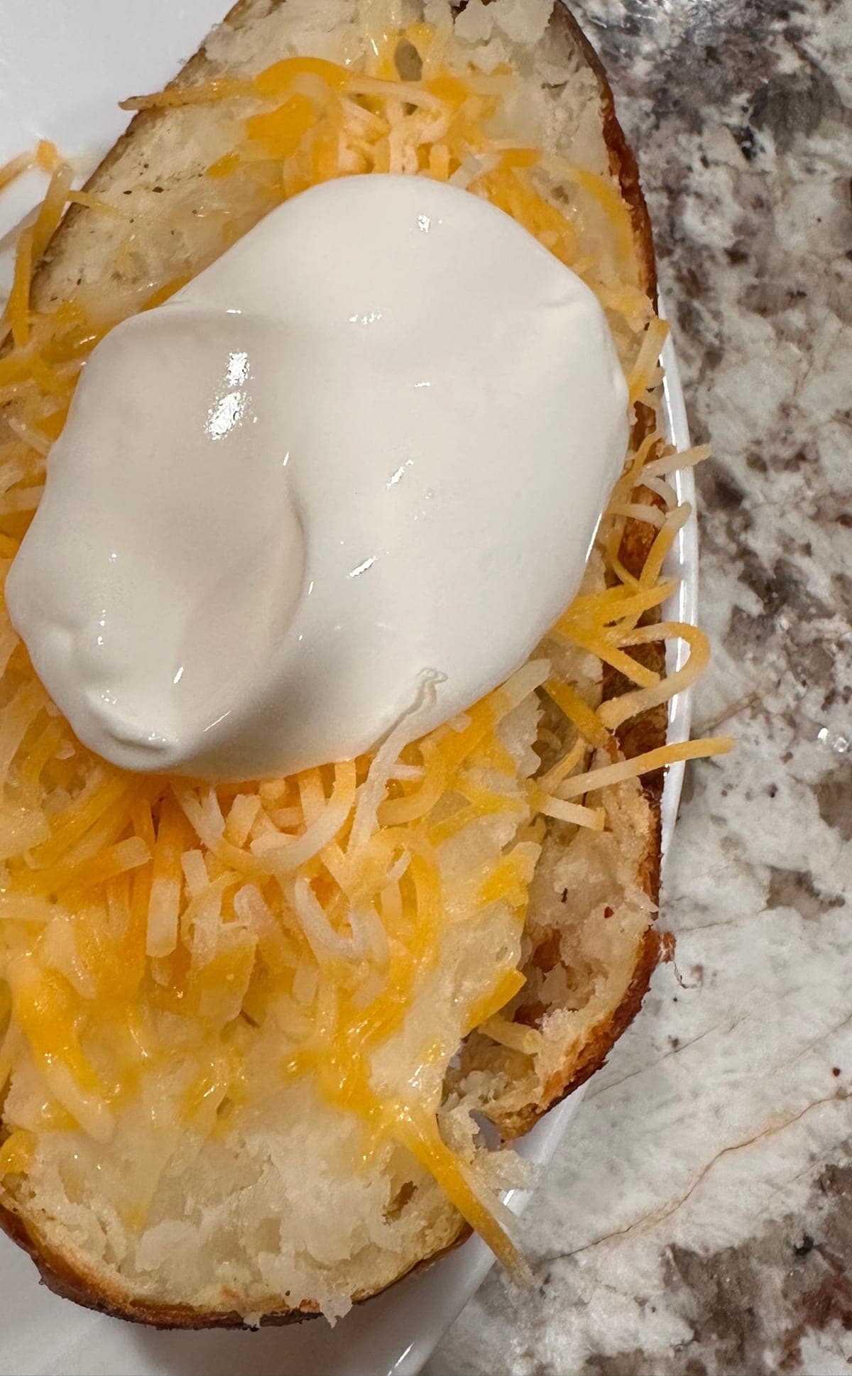 A baked potato split open on a white plate — the interior forked loose and steaming under a heap of shredded cheddar, with a spoonful of sour cream on top and the crisp brown skin curling back at the edges.