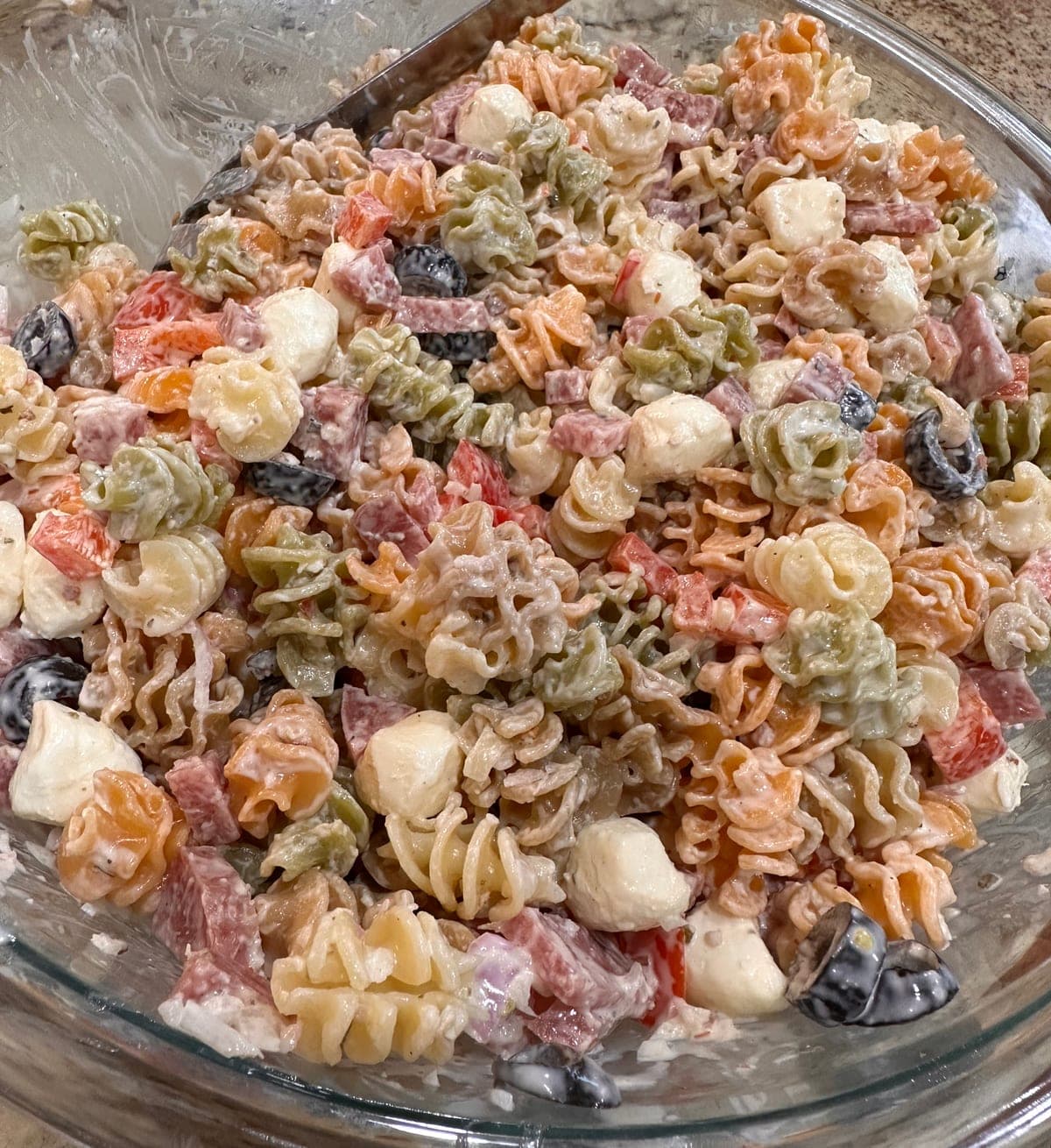 A glass bowl of creamy Italian pasta salad seen from above: tricolour pasta coated in dressing, with diced salami, mozzarella, red bell pepper and black olives, and a serving spoon resting at the back left.