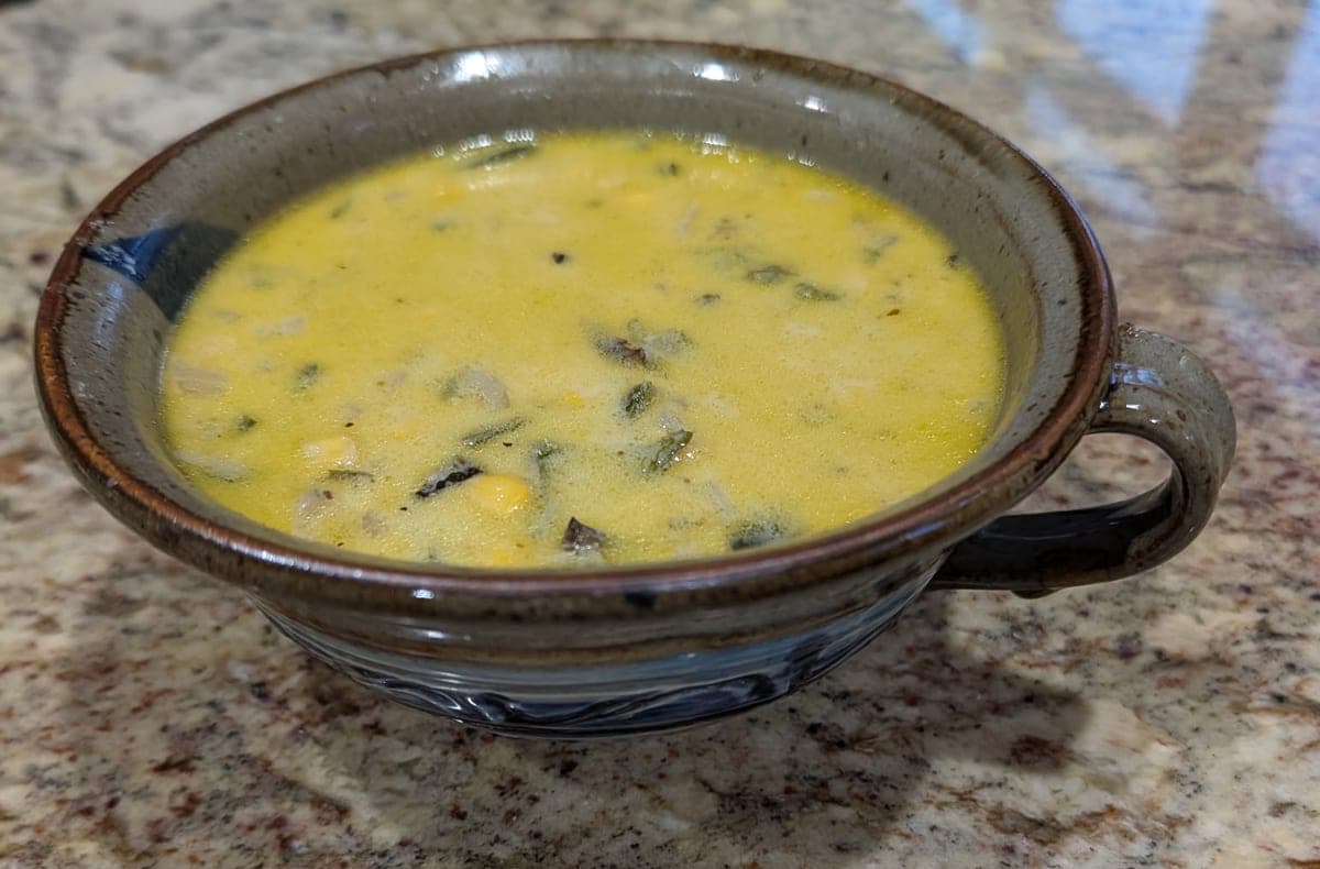 A handled stoneware bowl of pale yellow poblano soup, flecked with pieces of charred poblano, corn kernels and onion, on a granite counter.