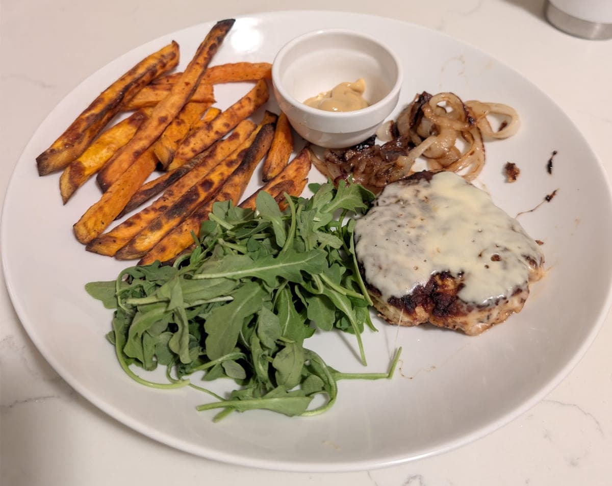 A white plate of roasted sweet potato fries beside a small cup of honey mustard, a tangle of caramelized onions, a chicken burger under melted cheddar and a pile of arugula.