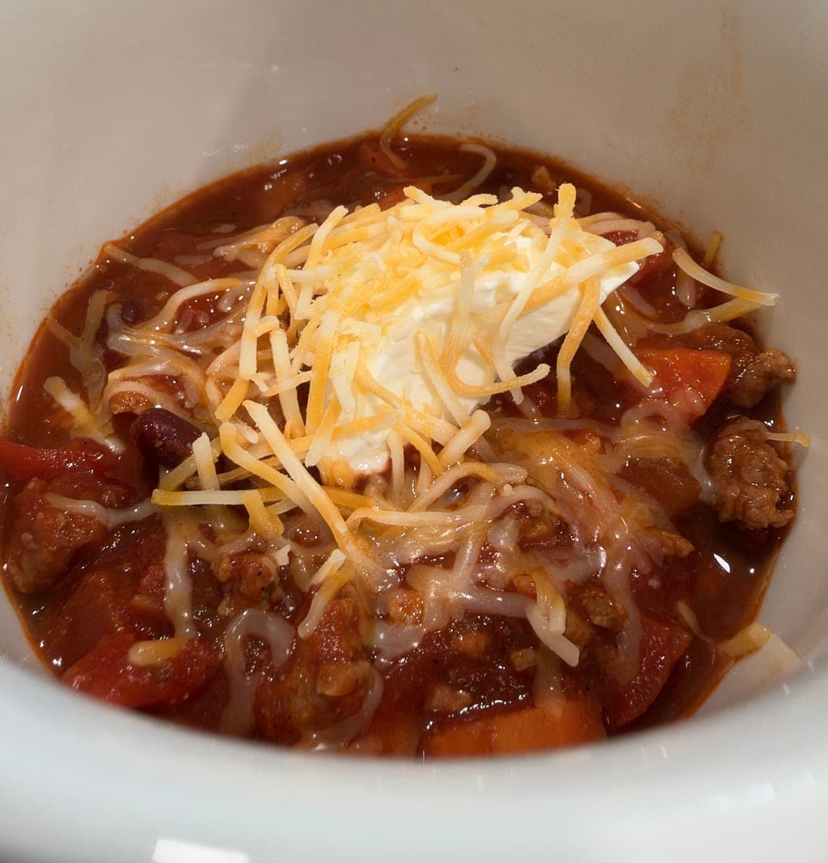 A white bowl of Italian sausage chili topped with a spoonful of sour cream and shredded cheese, with chunks of sausage, carrot, diced tomato and kidney beans in a thick red sauce.
