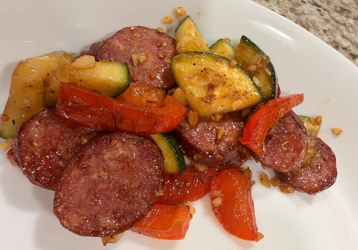 A white plate of browned kielbasa coins tossed with seared zucchini half-moons and strips of red bell pepper, flecked with golden minced garlic.