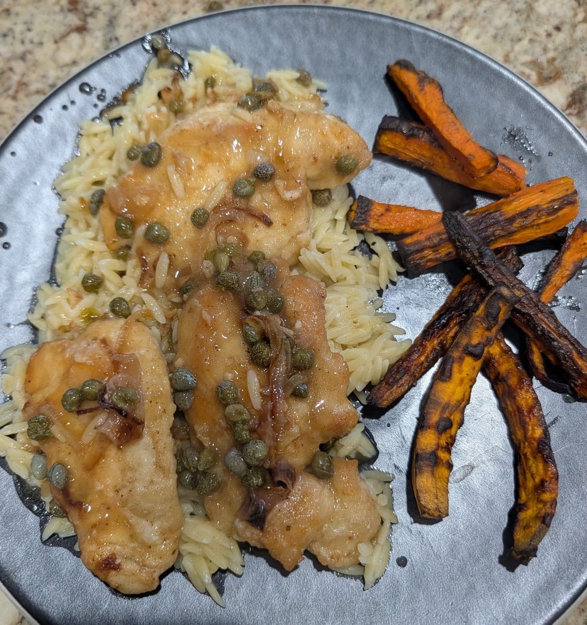 A gray plate of seared chicken cutlets over orzo, glossy with pan sauce and scattered with capers and sliced shallot, beside a pile of roasted carrot sticks.