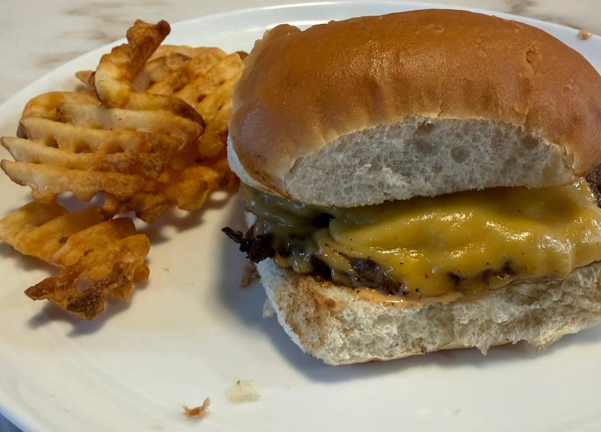 A white plate with a cheeseburger on a soft bun, a charred beef patty and crisp burnt onion under melted cheese with sauce on the bottom bun, beside a pile of waffle fries.