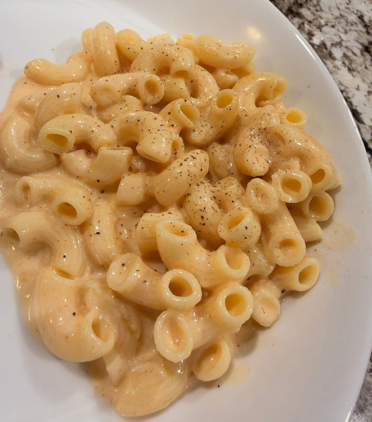 A wide white bowl of elbow macaroni in a smooth, pale orange cheese sauce, dusted with black pepper and flecks of paprika.