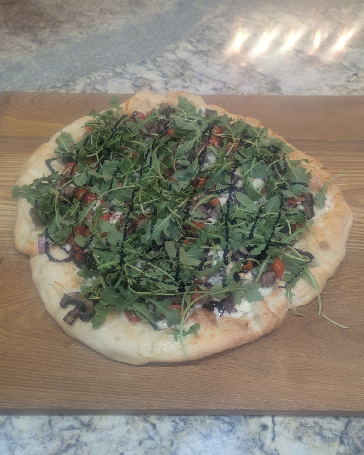 A pizza on a wooden board, heaped with arugula and zigzagged with balsamic glaze over cherry tomatoes, steak and blue cheese, with a few mushroom slices showing at the edge of the crust.