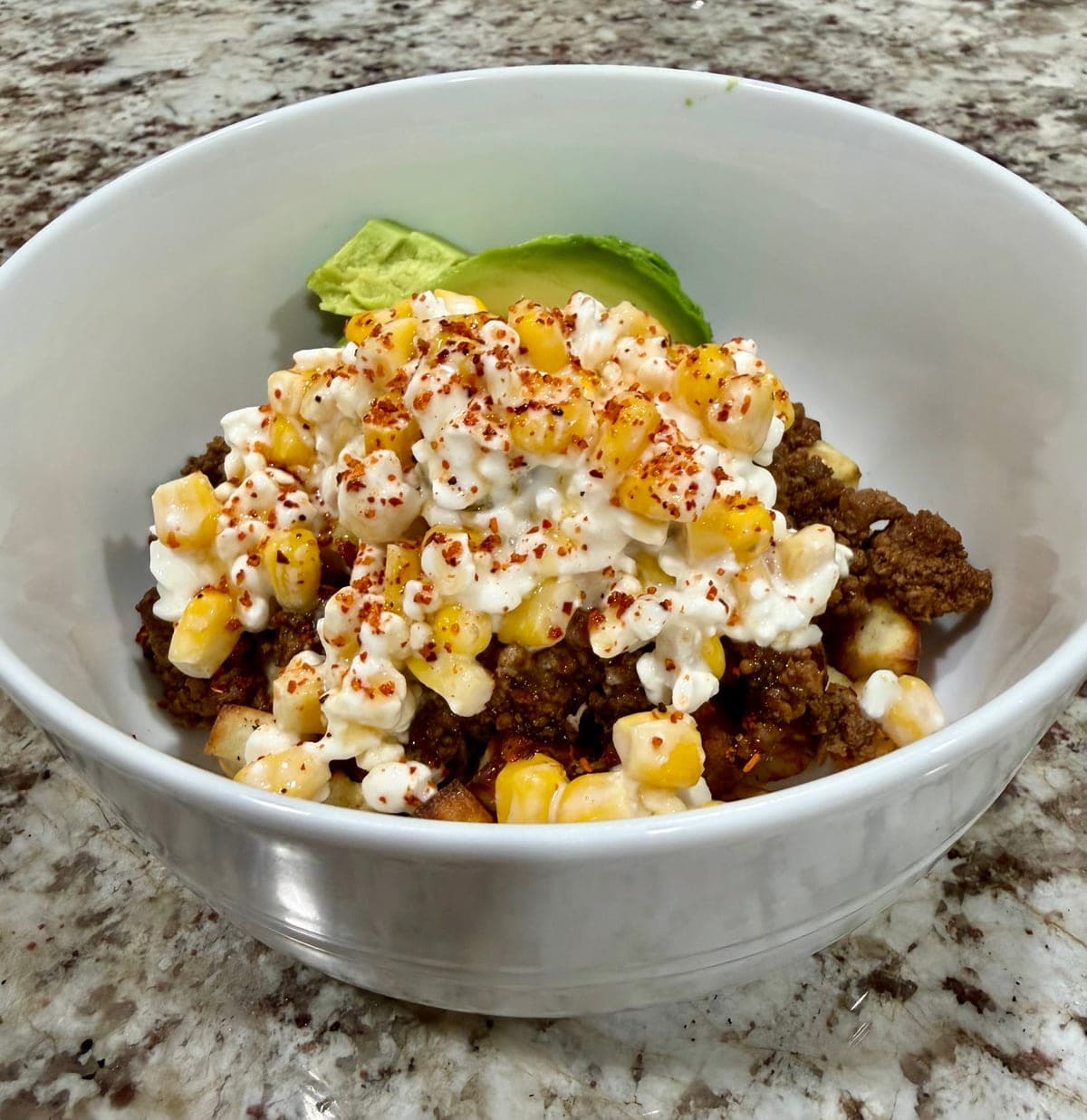 A white bowl of cubed roasted sweet potato and taco-seasoned ground beef under a heap of creamy corn and cottage cheese salsa, dusted with red Tajín and glossed with hot honey, two slices of avocado tucked in at the back.